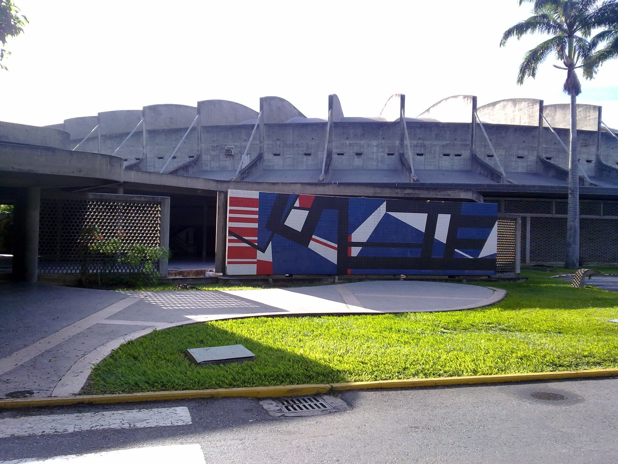Scalloped concrete roofline of the Aula Magna at the Ciudad Universitaria, with a tiled mural in front and a royal palm
