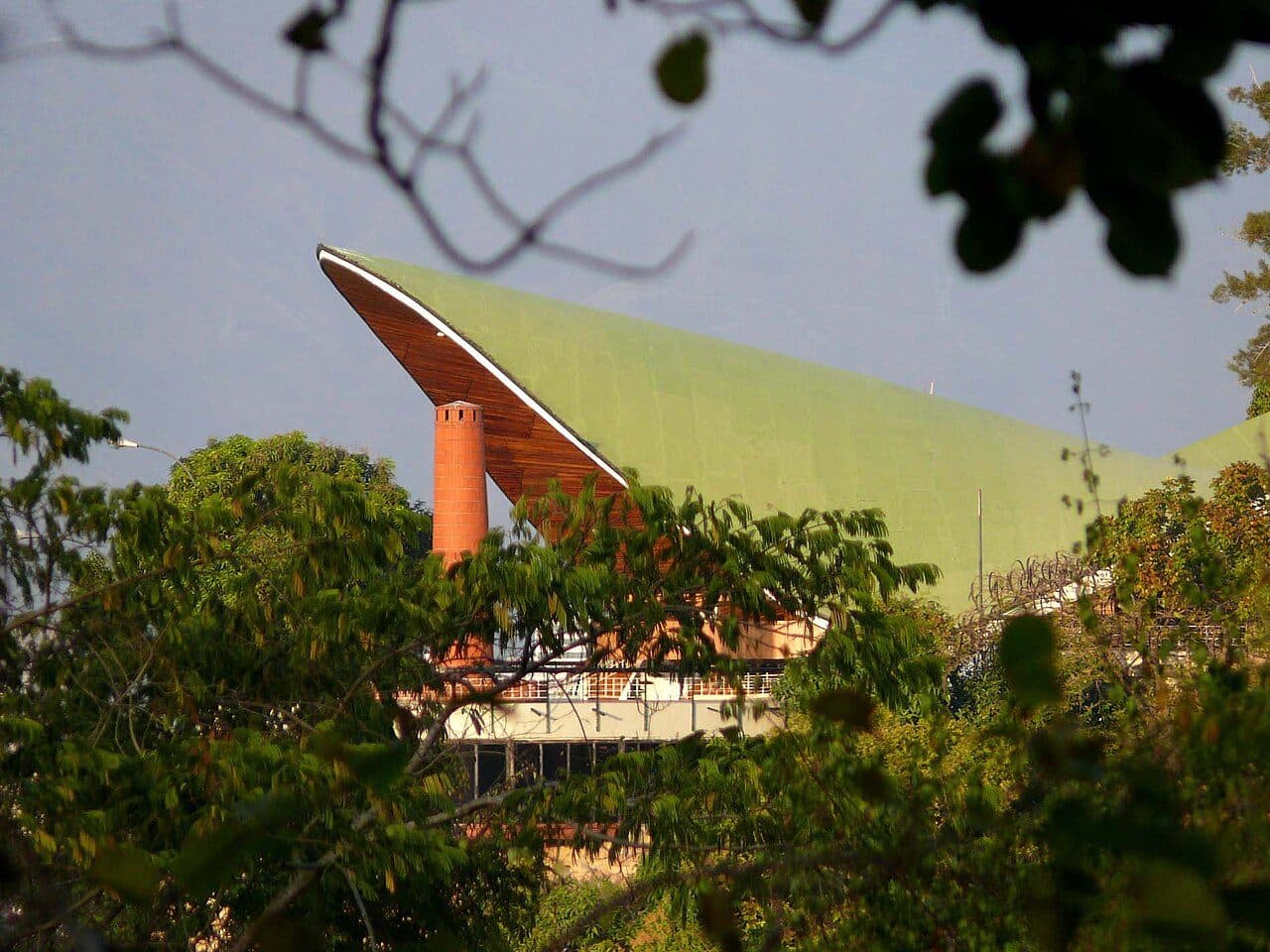 Curved green roof and brick chimney of the Club Táchira seen through trees