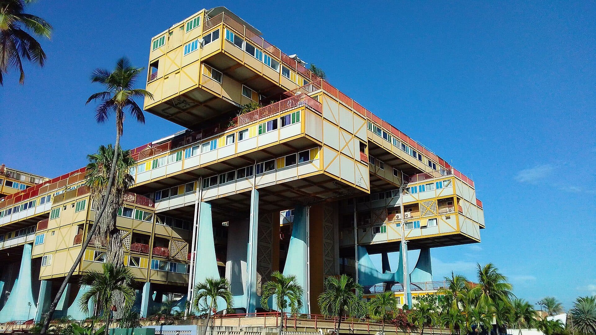 Raised yellow apartment blocks on pale blue pillars among palm trees, Edificio Árbol para Vivir