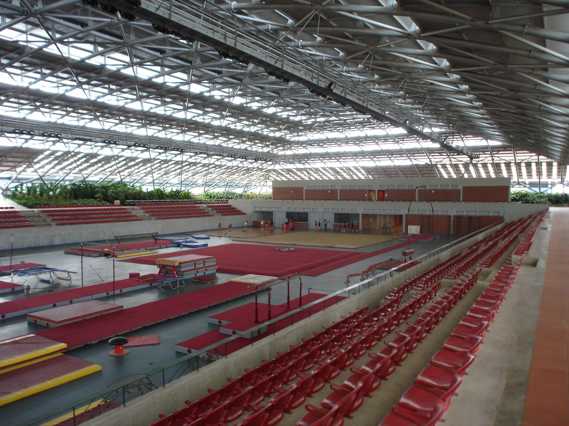 Indoor gymnastics hall with red seating under a lattice steel roof and planting along the far wall