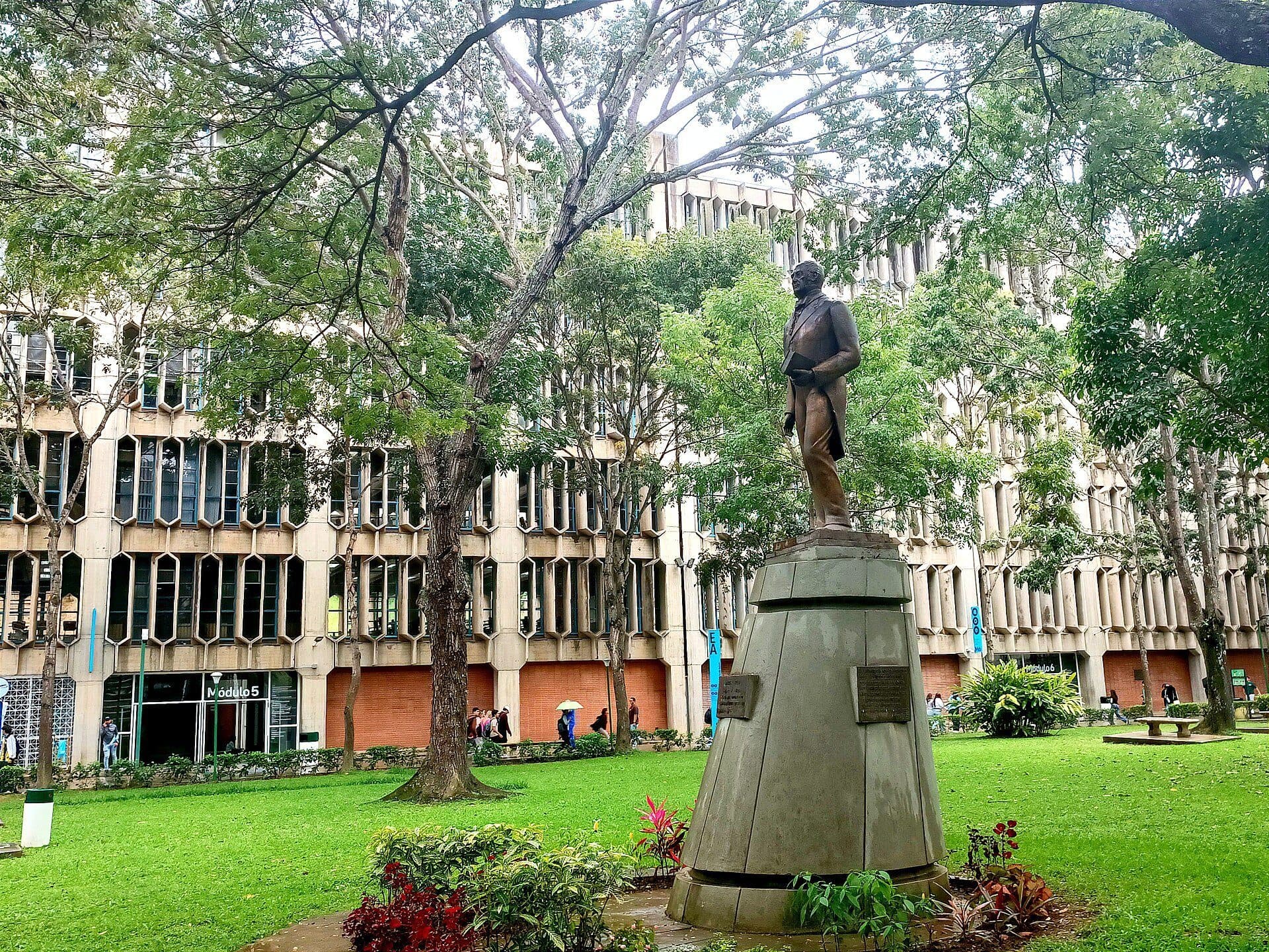 Hexagonal-windowed concrete classroom blocks on the Montalbán campus of Universidad Católica Andrés Bello, seen across a lawn with the Andrés Bello statue