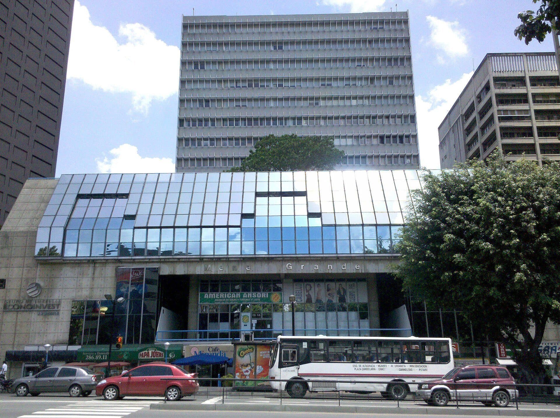Glass-and-concrete commercial annex of the Edificio Mene Grande in Caracas, with the office tower rising behind and a mature tree in front